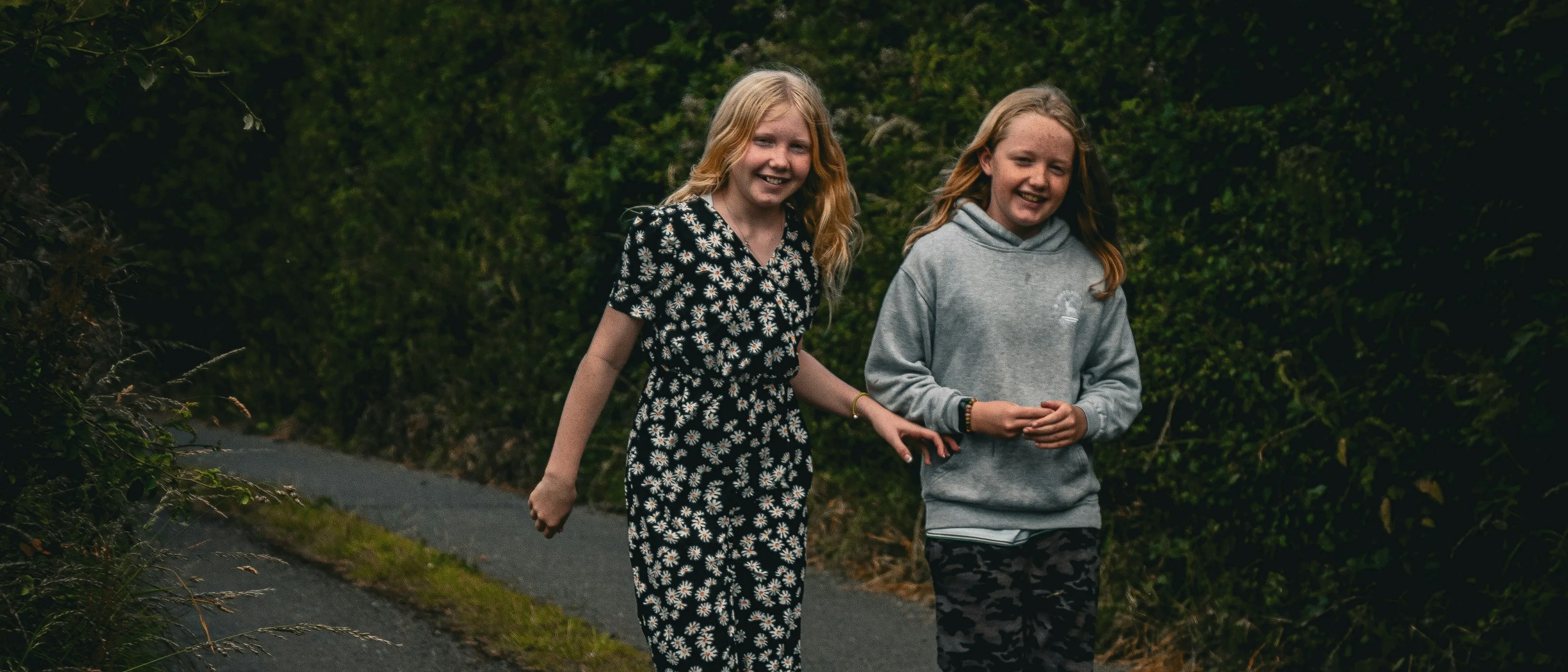 Zwei Mädchen gehen an einem Sommertag nebeneinander einen schmalen, von hohen Hecken gesäumten Weg in der Grafschaft Tipperary entlang und lächeln in die Kamera, die eine im geblümten Overall, die andere im grauen Kapuzenpullover.