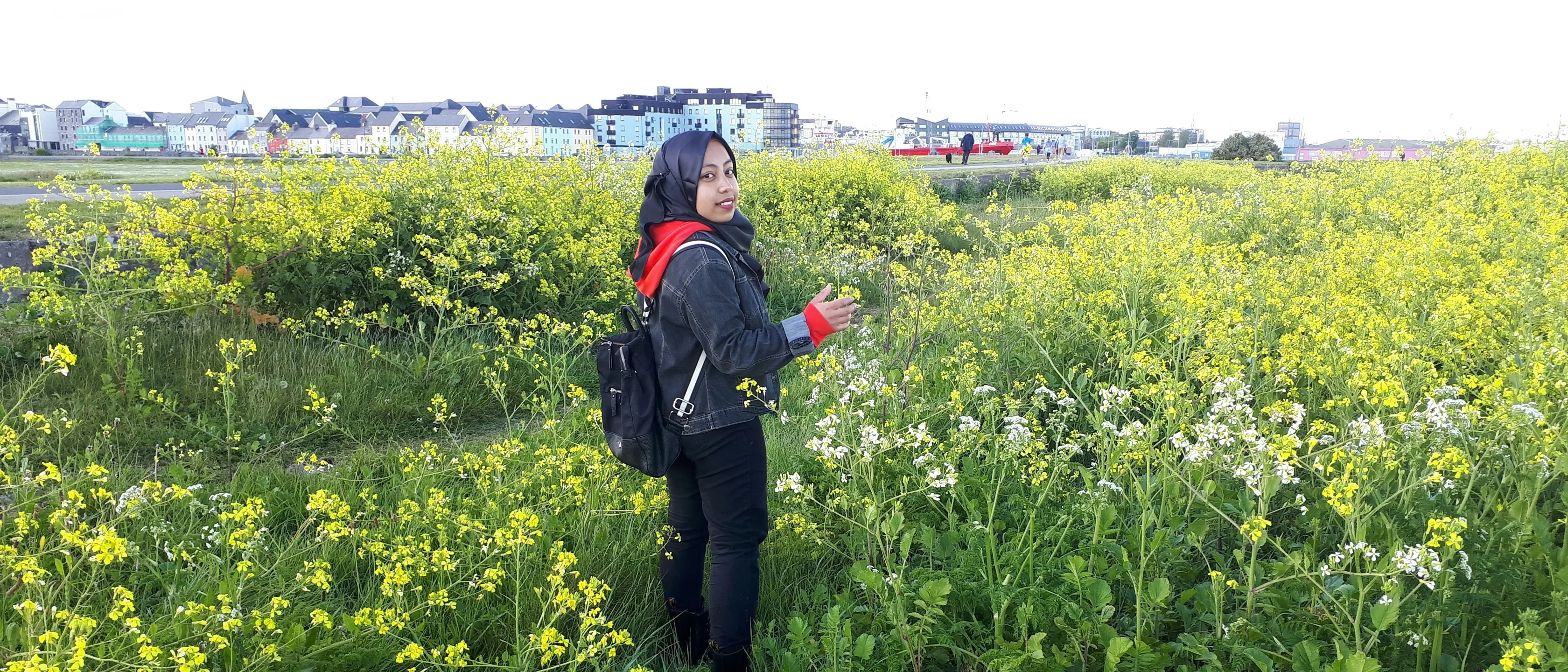 Eine junge Frau mit Kopftuch und Jeansjacke steht in einer Wiese voller gelber und weißer Wildblumen in Salthill bei Galway und lächelt über die Schulter in die Kamera, dahinter bunte Häuserreihen am Wasser.
