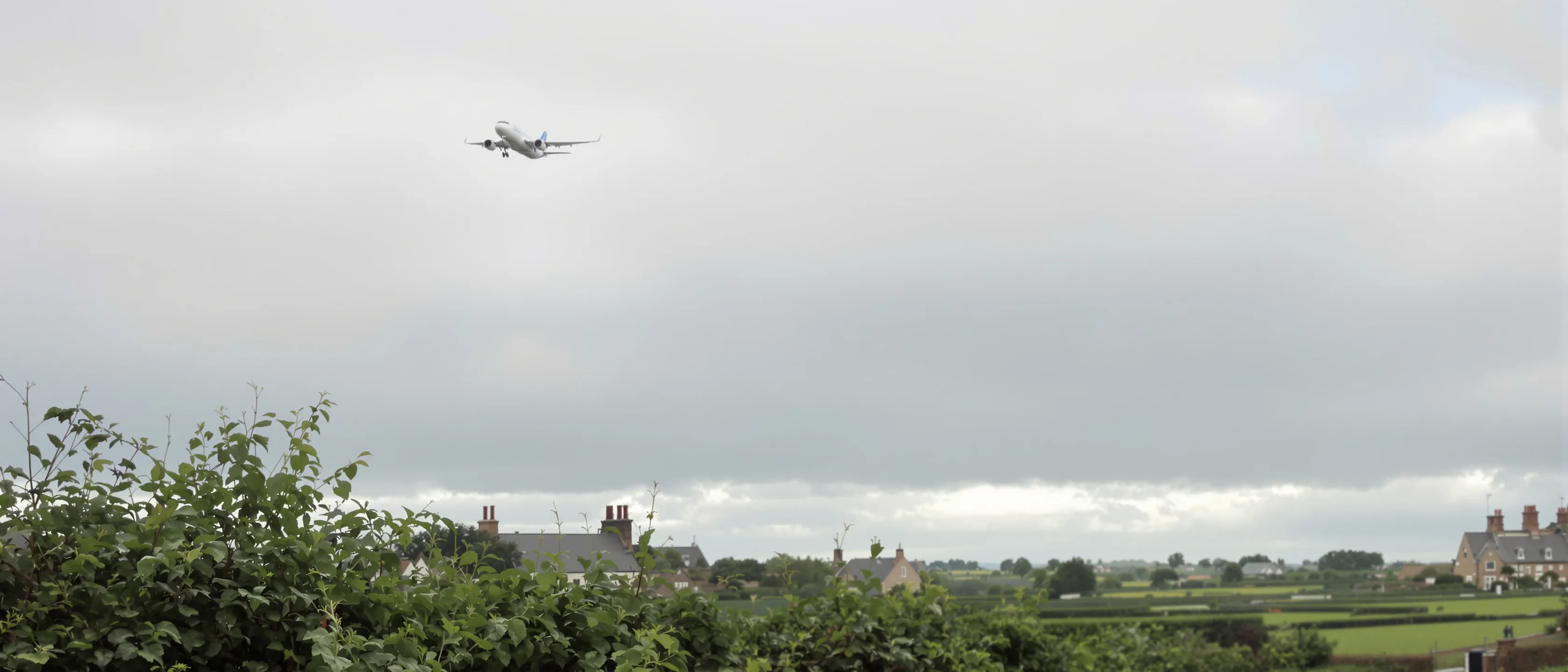 Ein Verkehrsflugzeug steigt klein vor einem weiten grauen Wolkenhimmel auf, darunter eine Hecke, Hausdächer und die grünen Felder der Umgebung.
