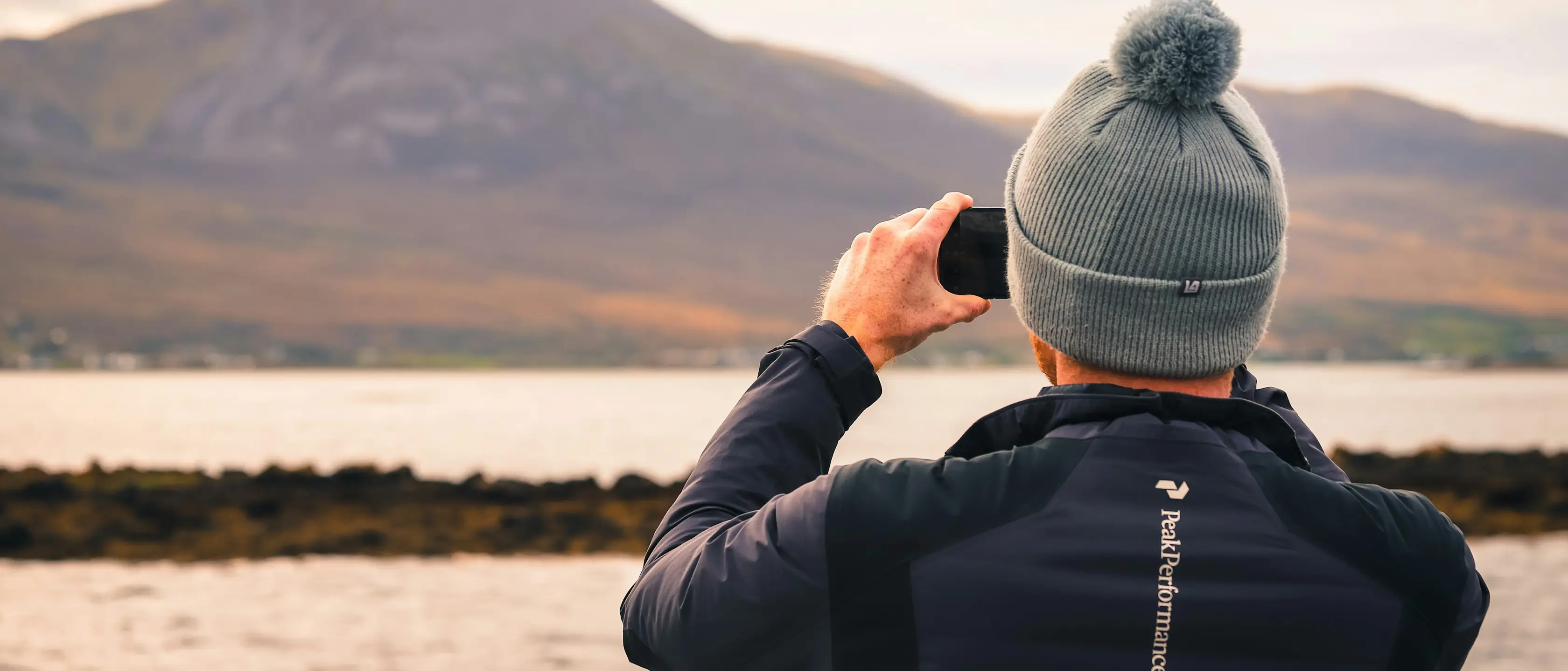 Ein Mensch mit Bommelmütze fotografiert eine Bucht mit dem Telefon.