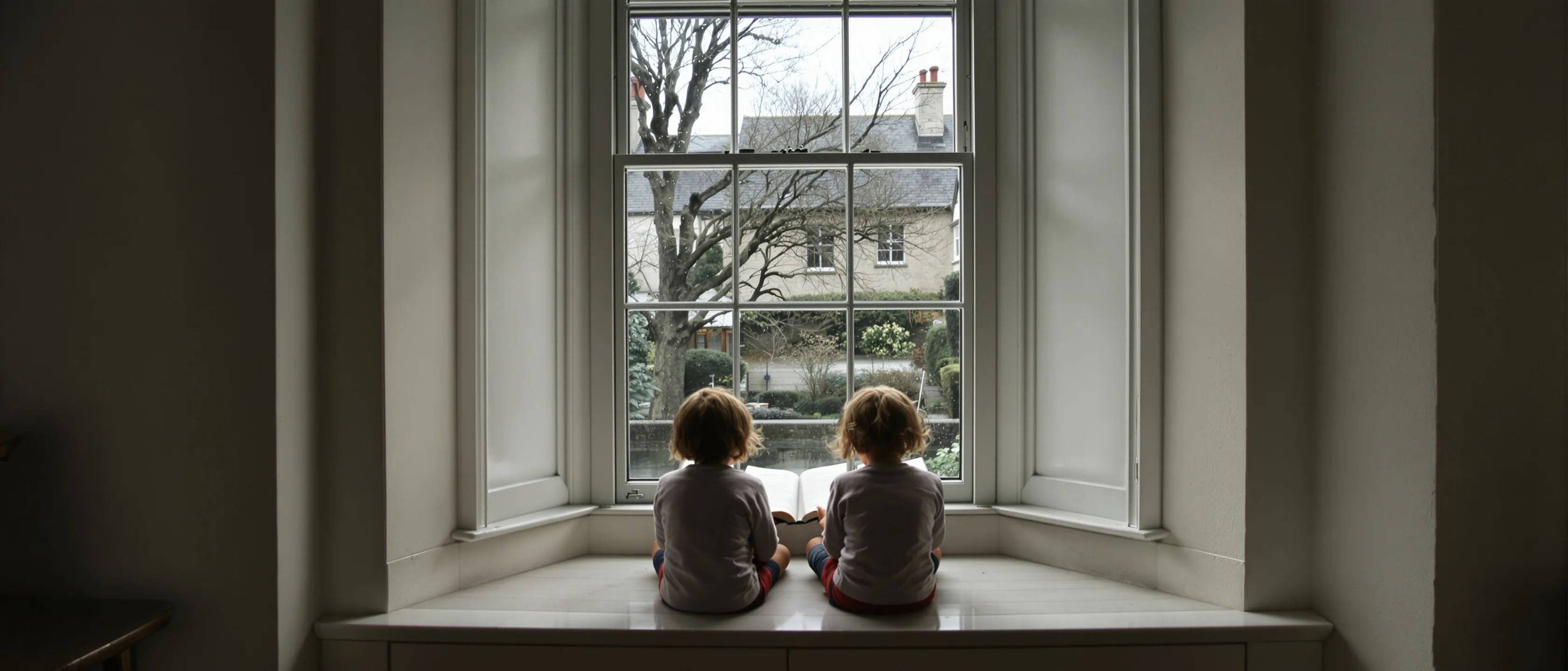 Zwei Kinder sitzen von hinten gesehen auf der breiten Fensterbank eines Sprossenfensters und lesen gemeinsam in einem aufgeschlagenen Buch; draußen ein winterlicher Garten.
