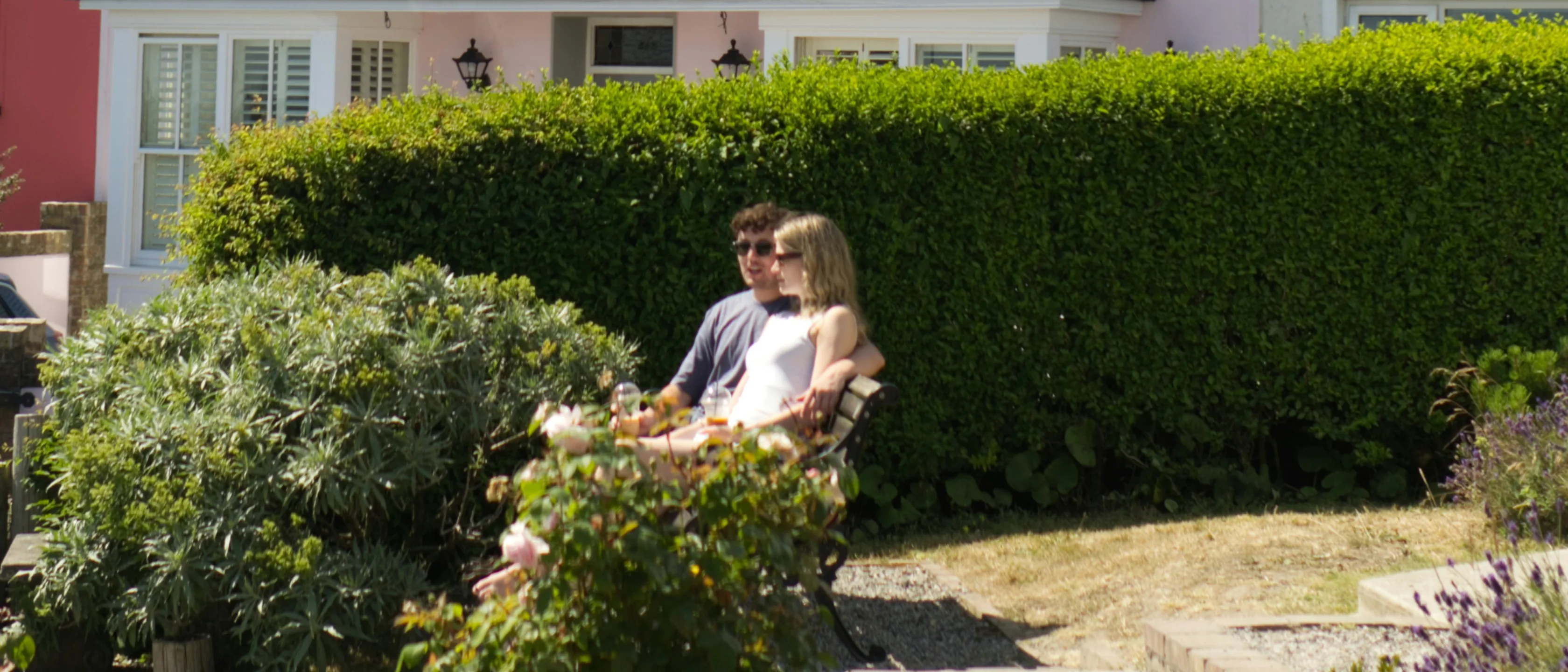 Ein junges Paar sitzt an einem Sommertag auf einer Bank in einem Vorgarten in Dunmore East, Grafschaft Waterford, vor einer hohen Hecke und einem rosa Reihenhaus.