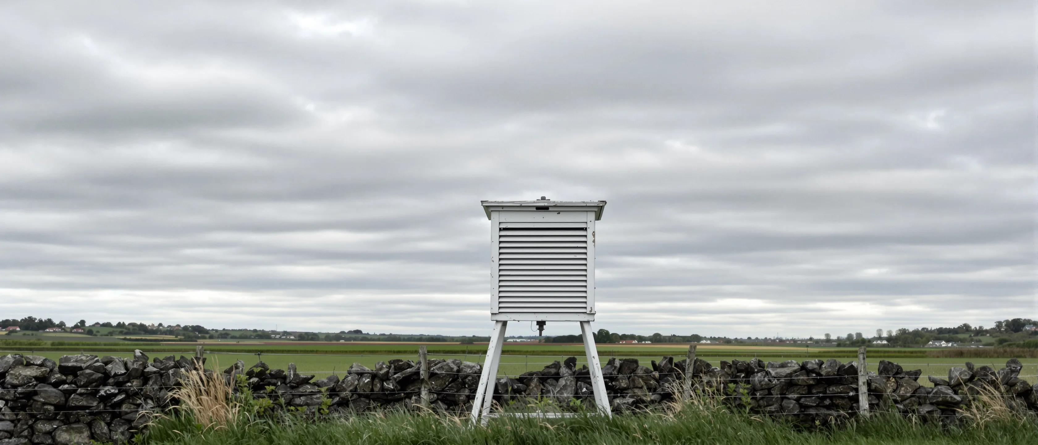 Eine weiße Wetterhütte auf Beinen steht auf einer gemähten Wiese hinter einer niedrigen Bruchsteinmauer, darüber ein weiter geschichteter grauer Himmel.