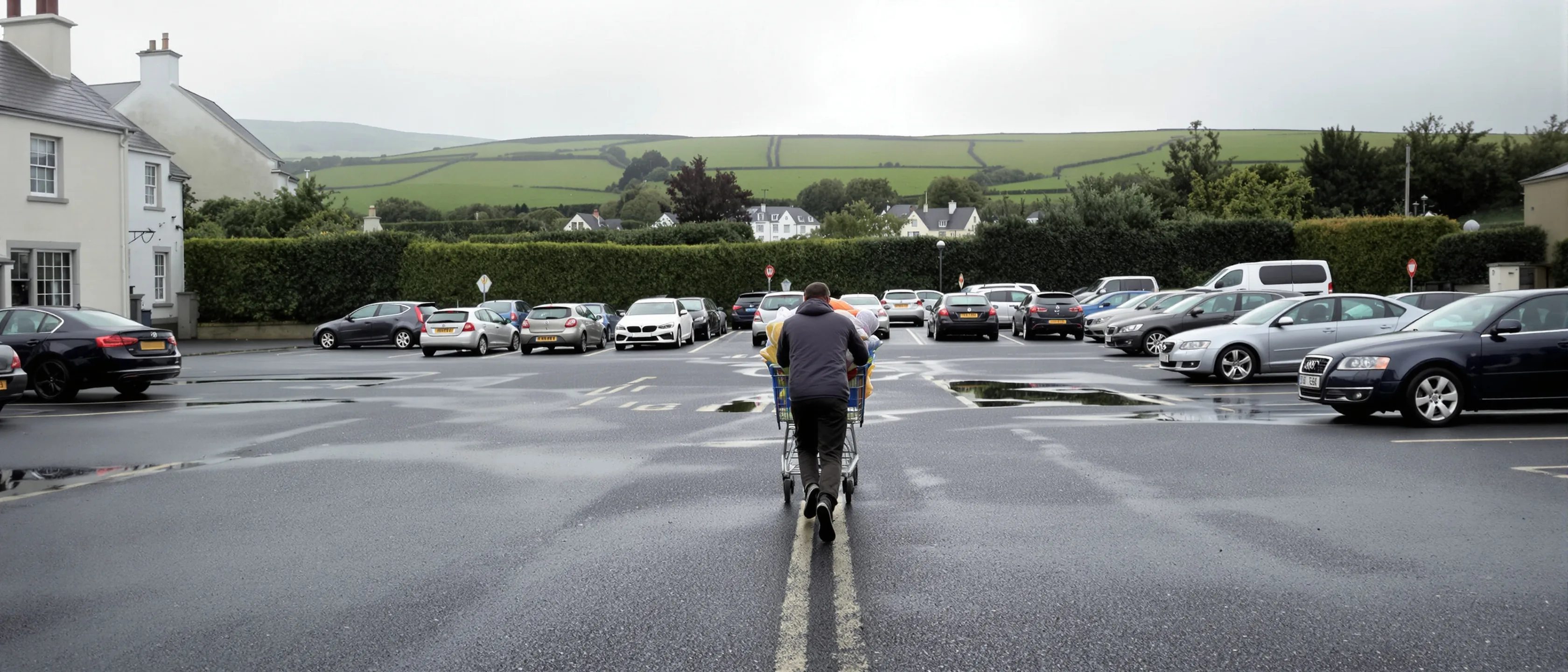 Ein Mensch schiebt von hinten gesehen einen vollen Einkaufswagen über einen nassen Parkplatz am Ortsrand; hinter den geparkten Autos beginnen sofort grüne Hecken und Felder.