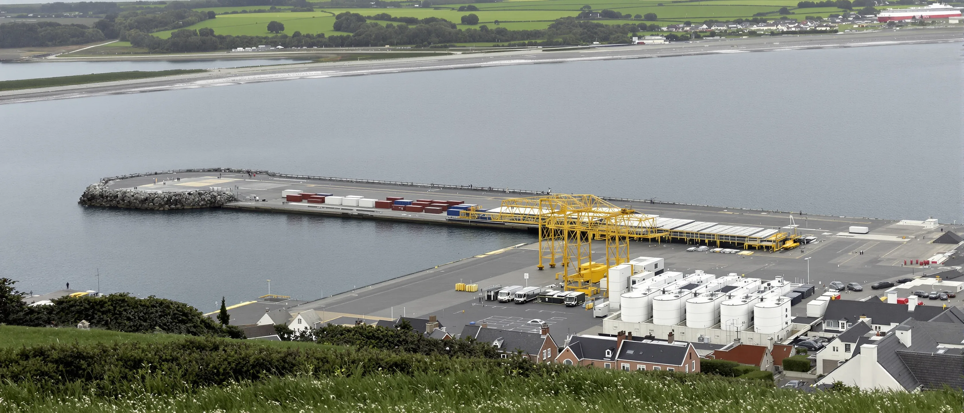 Blick von einer Anhöhe über einen Fährhafen: ein langer leerer Betonkai mit gelben Portalkränen, gestapelte Container, eine steinerne Mole und dahinter offenes graugrünes Wasser.
