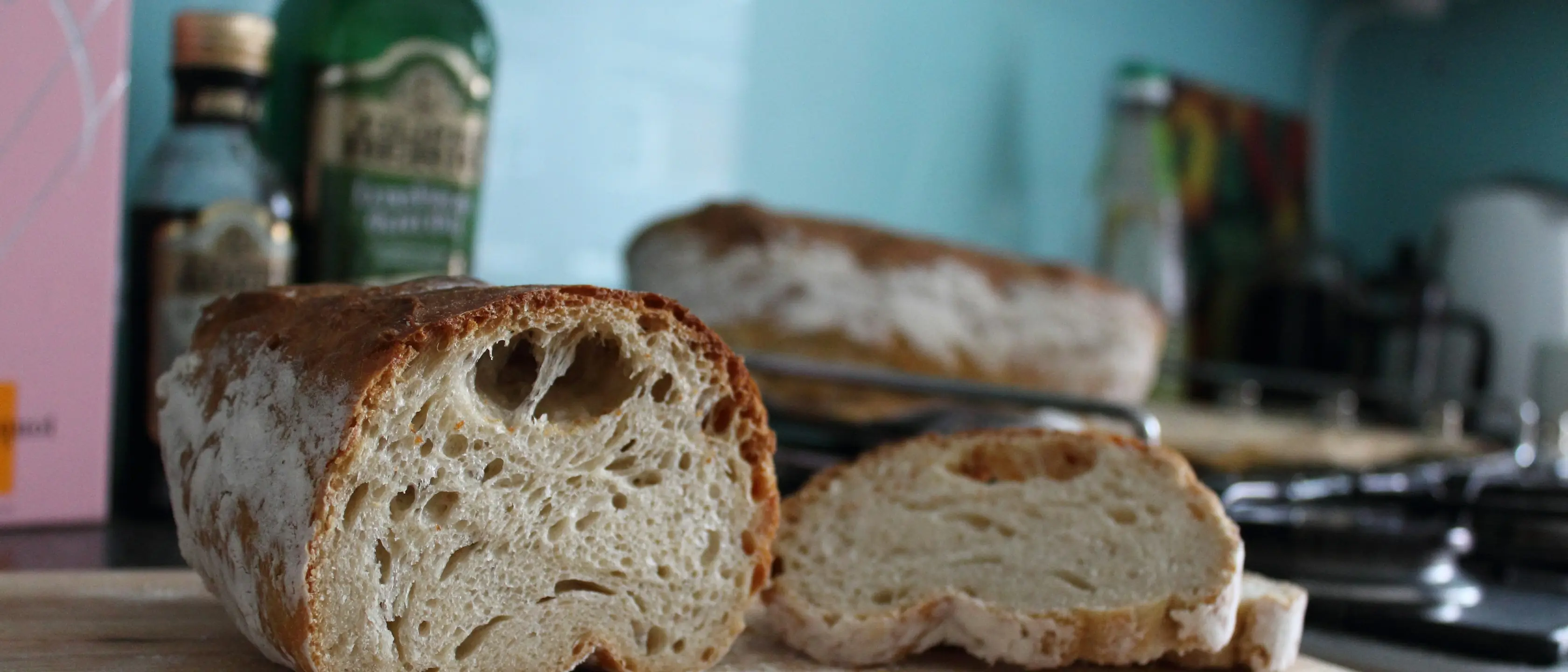 Aufgeschnittenes Brot auf einem Tisch, dahinter eine grüne Flasche und eine Küche.