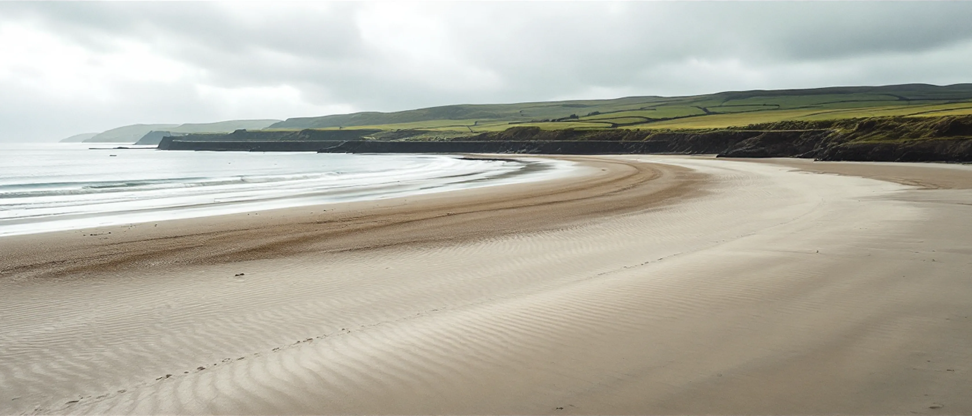 Eine weite Bucht bei Niedrigwasser, fester gerippter Sand, dahinter eine lange geschwungene Granit-Ufermauer und grünes Land.
