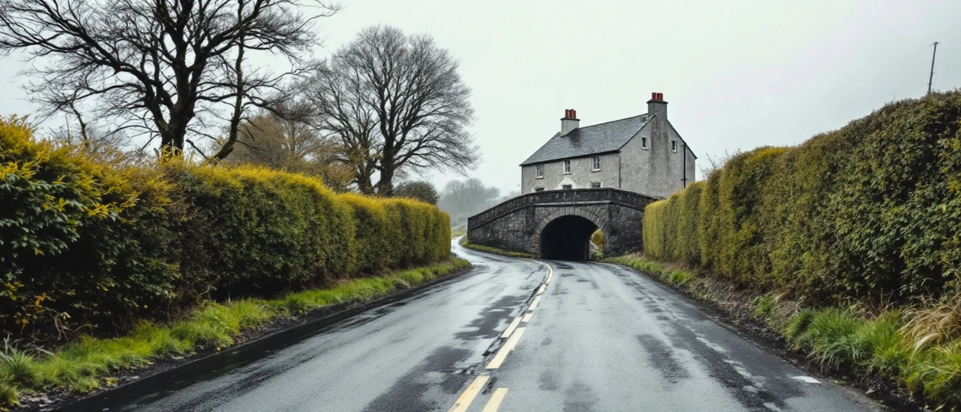 Eine schmale Landstraße führt zwischen hohen Hecken auf eine alte Steinbogenbrücke zu, dahinter ein graues Haus mit Schieferdach, nasser Asphalt.