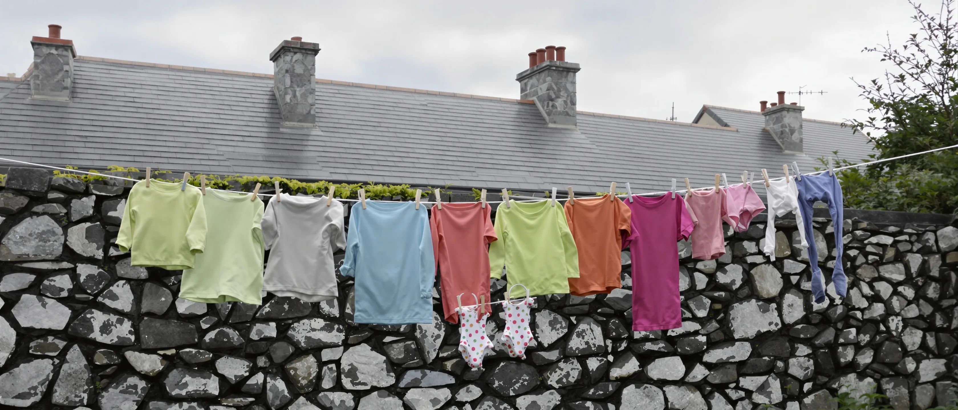 Eine Wäscheleine mit bunter Kinderkleidung vor einer Bruchsteinmauer, dahinter Schieferdächer und Schornsteine.