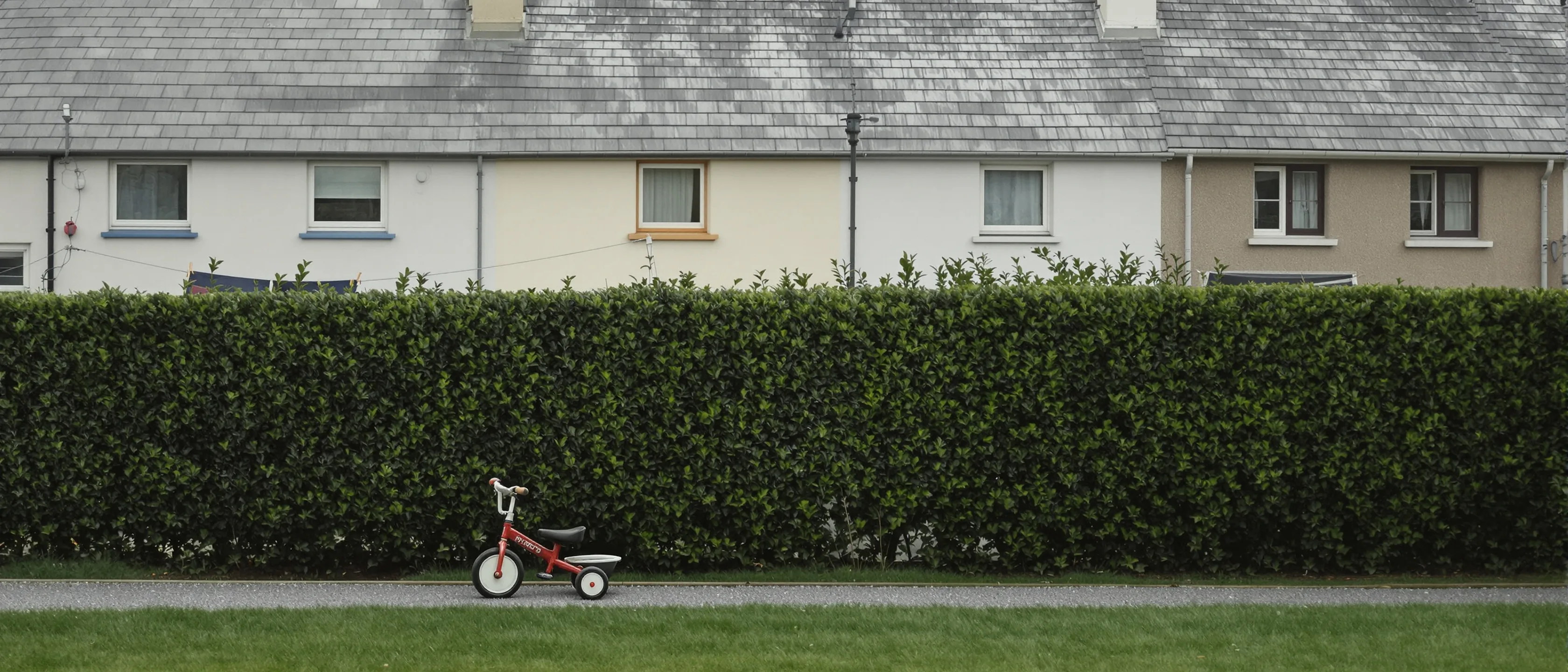 Ein rotes Dreirad auf einem Weg vor einer langen, geschnittenen Hecke, dahinter Obergeschosse und Schieferdächer einer Häuserzeile.