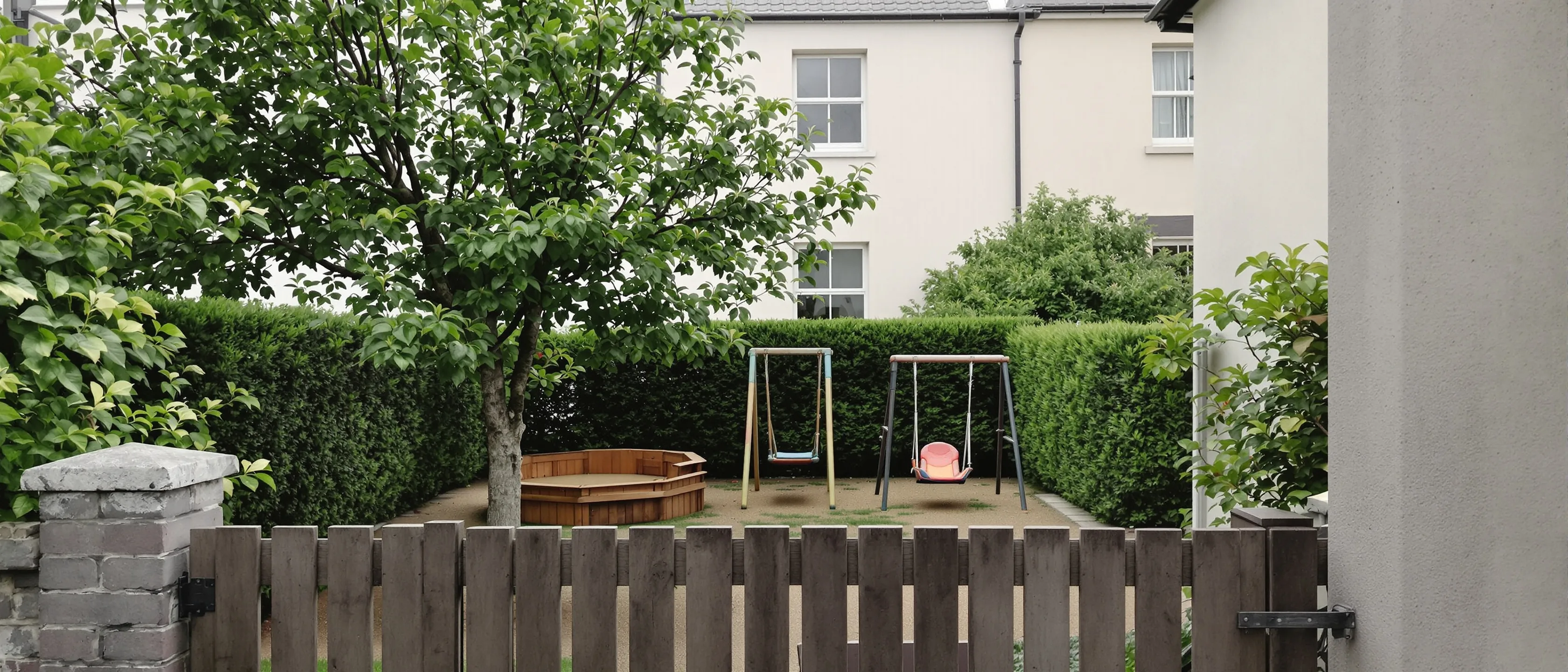 Ein Garten über einen Holzzaun gesehen: Schaukelgerüst unter einem Baum, dahinter ein helles Haus mit Sprossenfenstern.