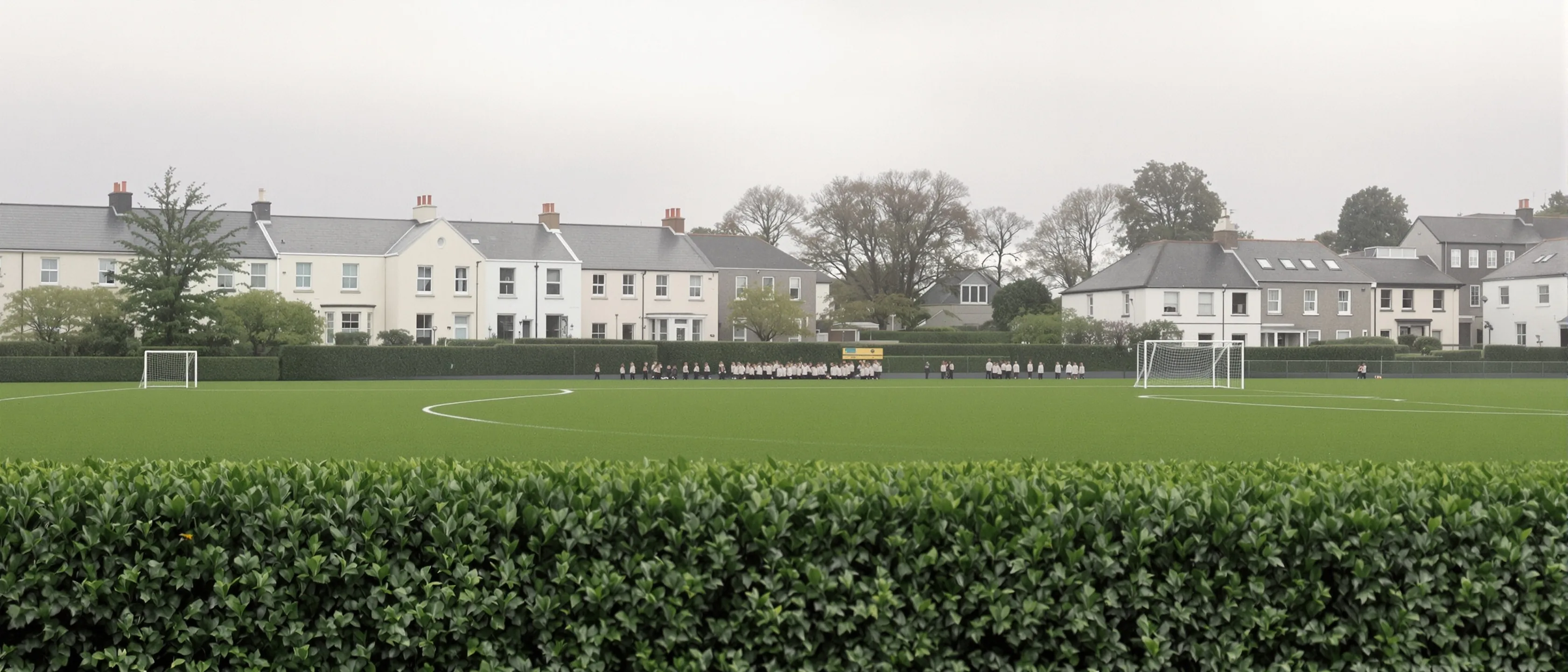 Ein weiter Sportplatz hinter einer Hecke, eine große Gruppe Kinder winzig in der Ferne, dahinter helle Häuser unter grauem Himmel.
