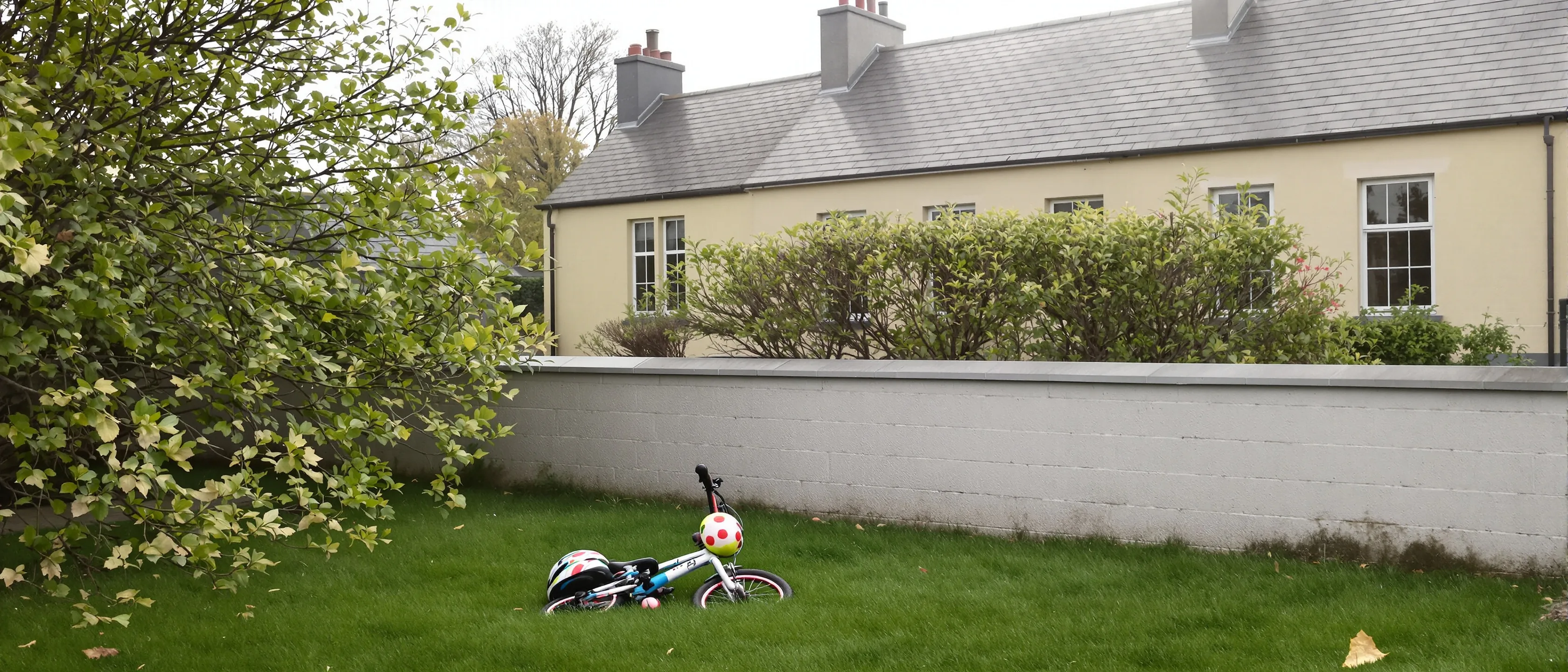 Ein Kinderfahrrad mit Helm liegt im Gras vor einer weißen Gartenmauer, dahinter ein langes cremefarbenes Gebäude mit Schieferdach unter Bäumen.