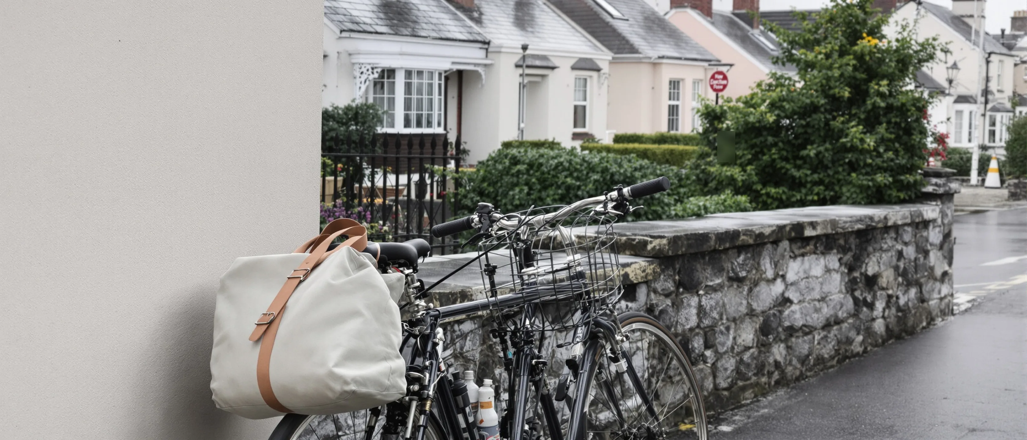 Ein Fahrrad mit heller Segeltuchtasche auf dem Gepäckträger lehnt an einem Steinpfeiler, dahinter eine ruhige Straße mit hellen Häusern.
