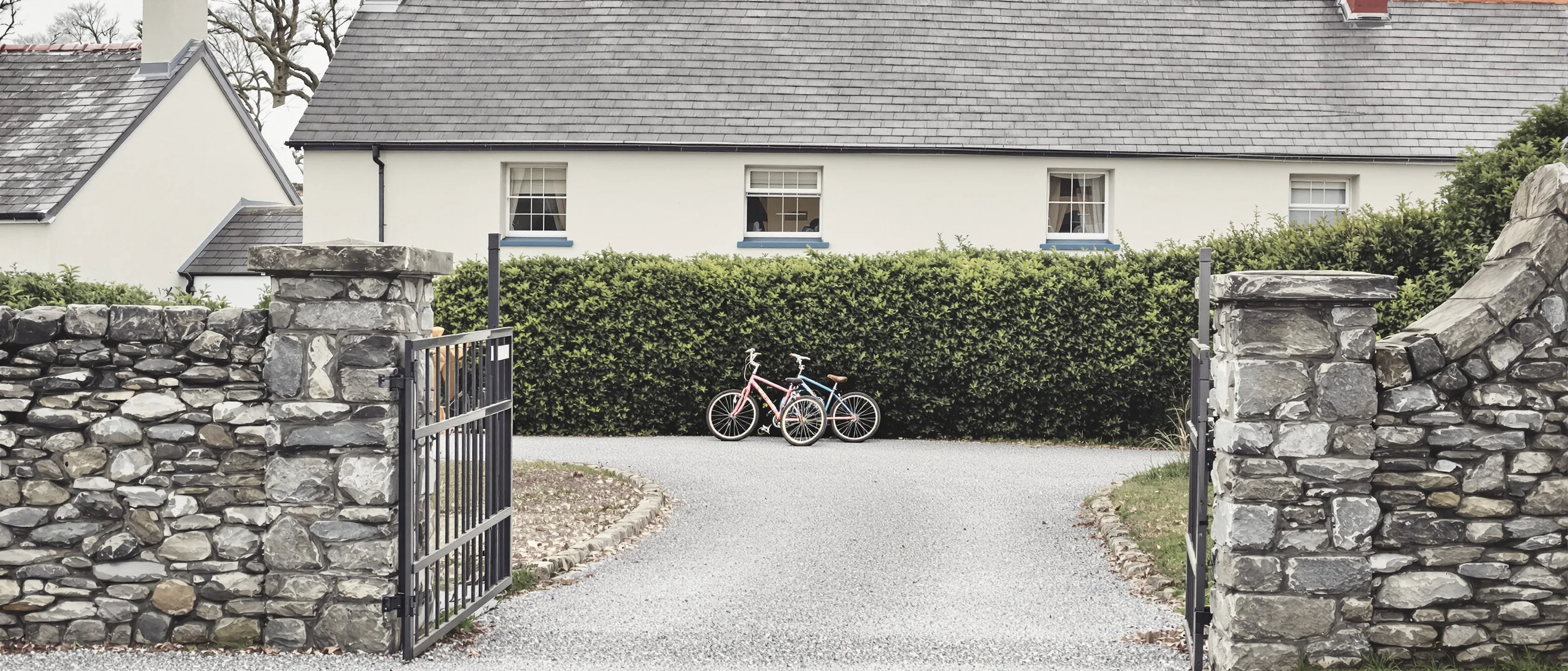 Eine Kieszufahrt zwischen Steinpfeilern, zwei Kinderfahrräder lehnen an der hohen Hecke, dahinter Obergeschoss und Schieferdach eines Hauses.