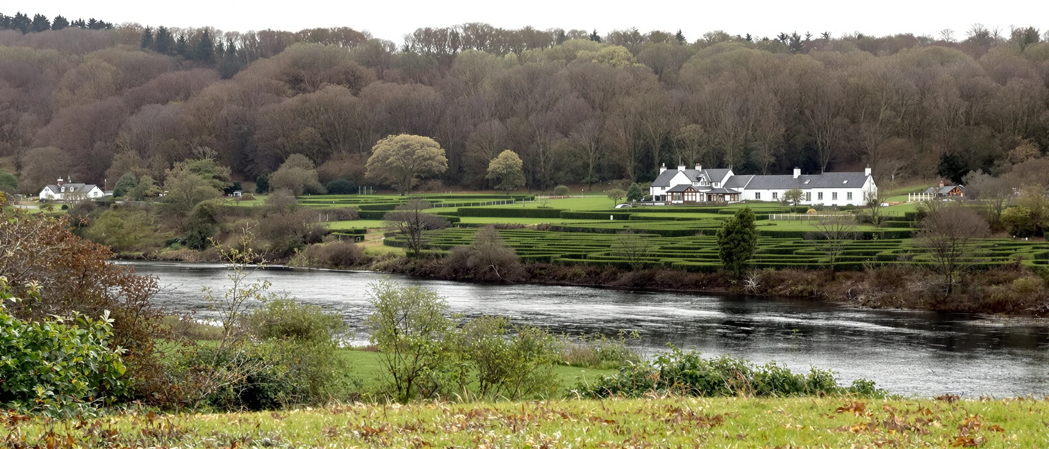 Ein breites bewaldetes Flusstal: der Fluss zieht ruhig zwischen alten Bäumen, am anderen Ufer Heckengärten und ein weißes Landhaus.