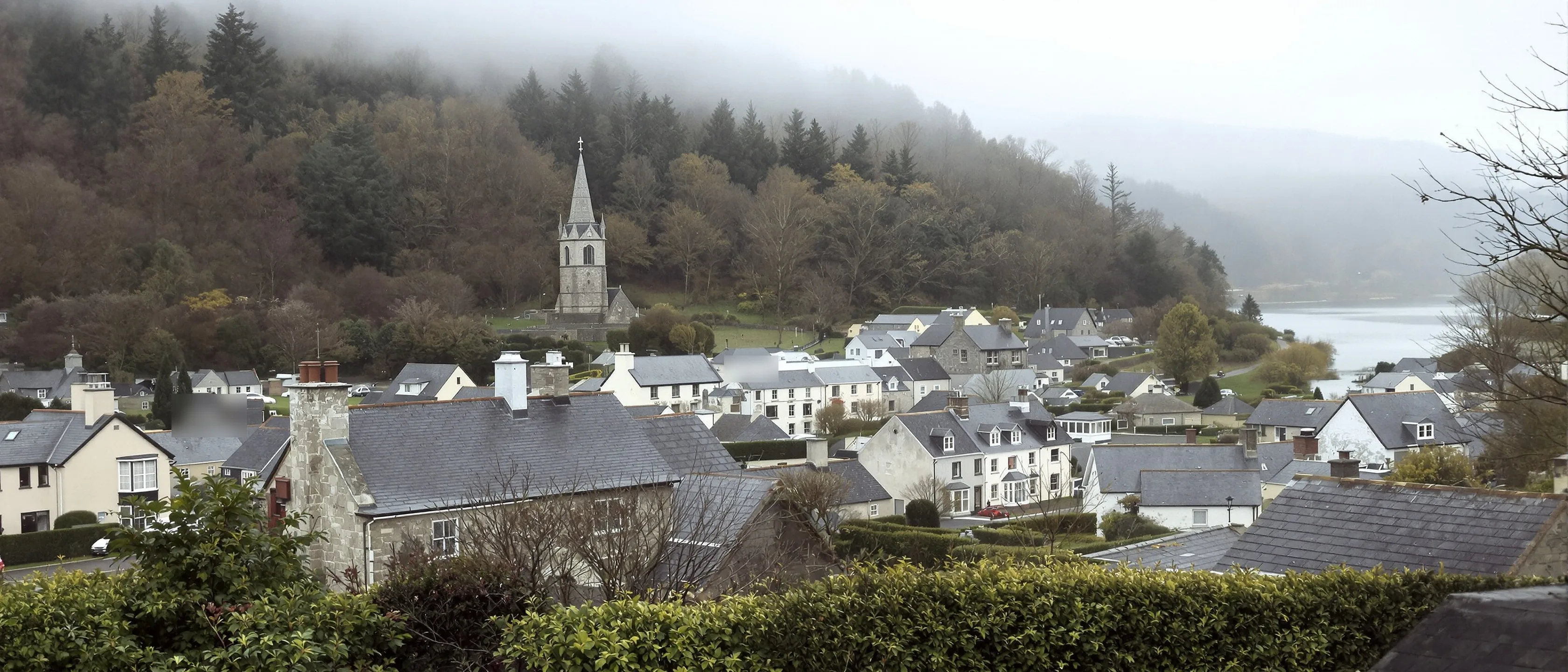 Ein kleiner Ort mit Schieferdächern und einem Kirchturm tief in einem bewaldeten Flusstal, Nebel hebt sich vom Wasser.