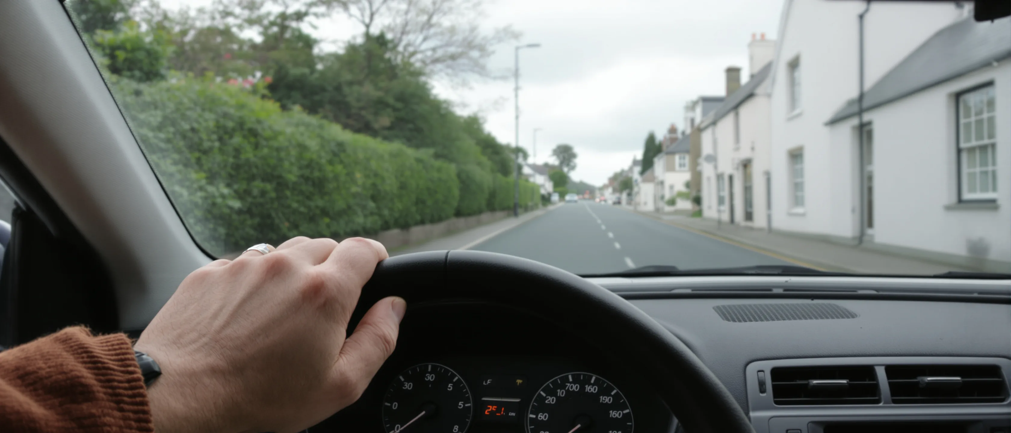 Hände am Lenkrad eines rechtsgelenkten Autos, Blick durch die Scheibe auf eine Dorfstraße, auf der links gefahren wird.