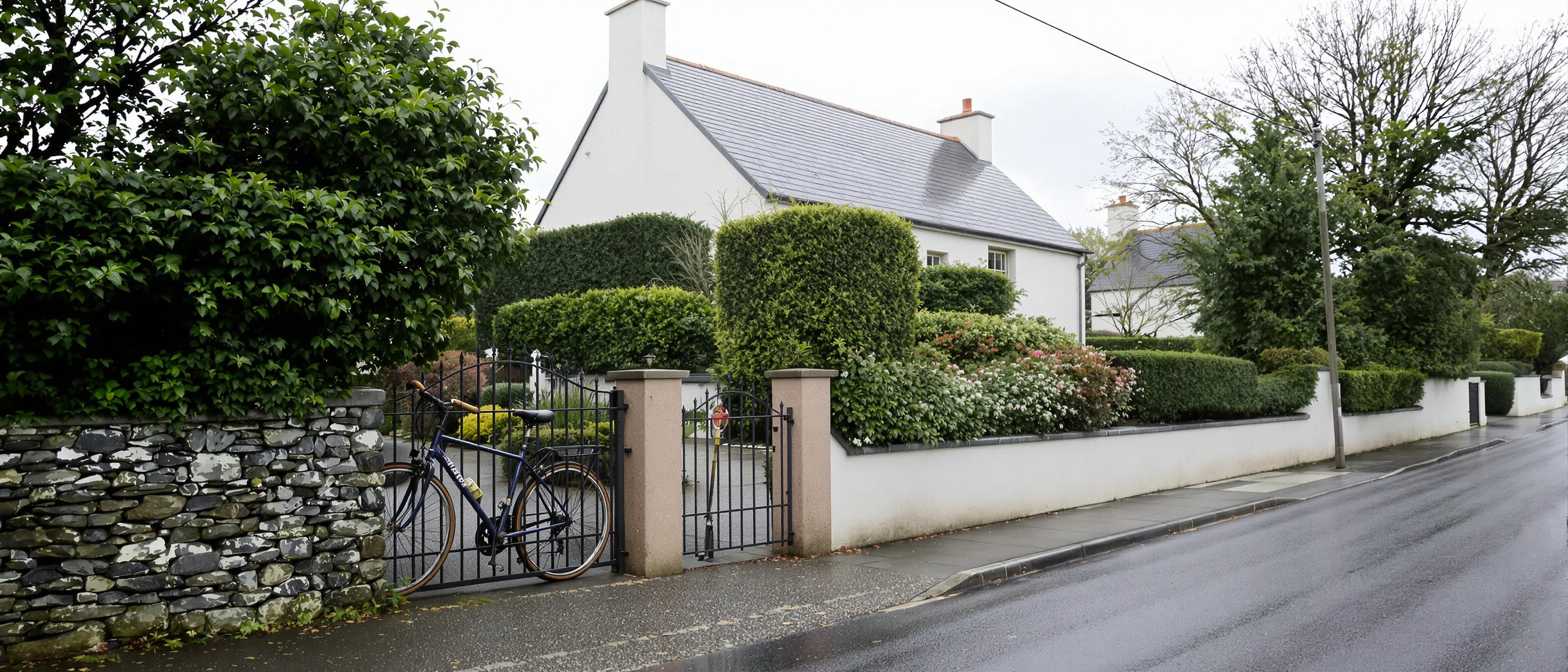 Einzelnes Haus hinter Gartenmauer und geschnittener Hecke an einer Dorfstraße, ein Fahrrad lehnt innen am Tor.