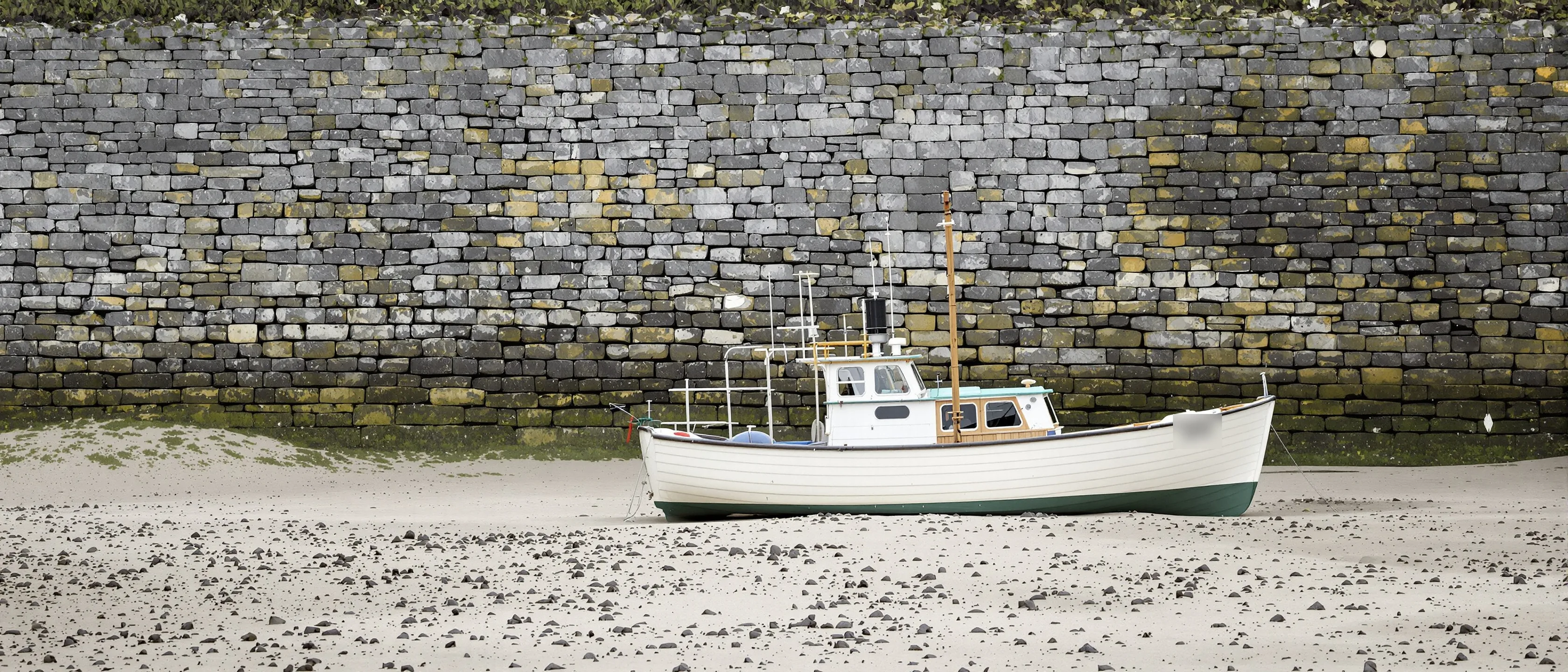 Ein kleines hölzernes Fischerboot steht aufrecht auf festem Sand bei Niedrigwasser, dahinter eine hohe Bruchsteinmauer.