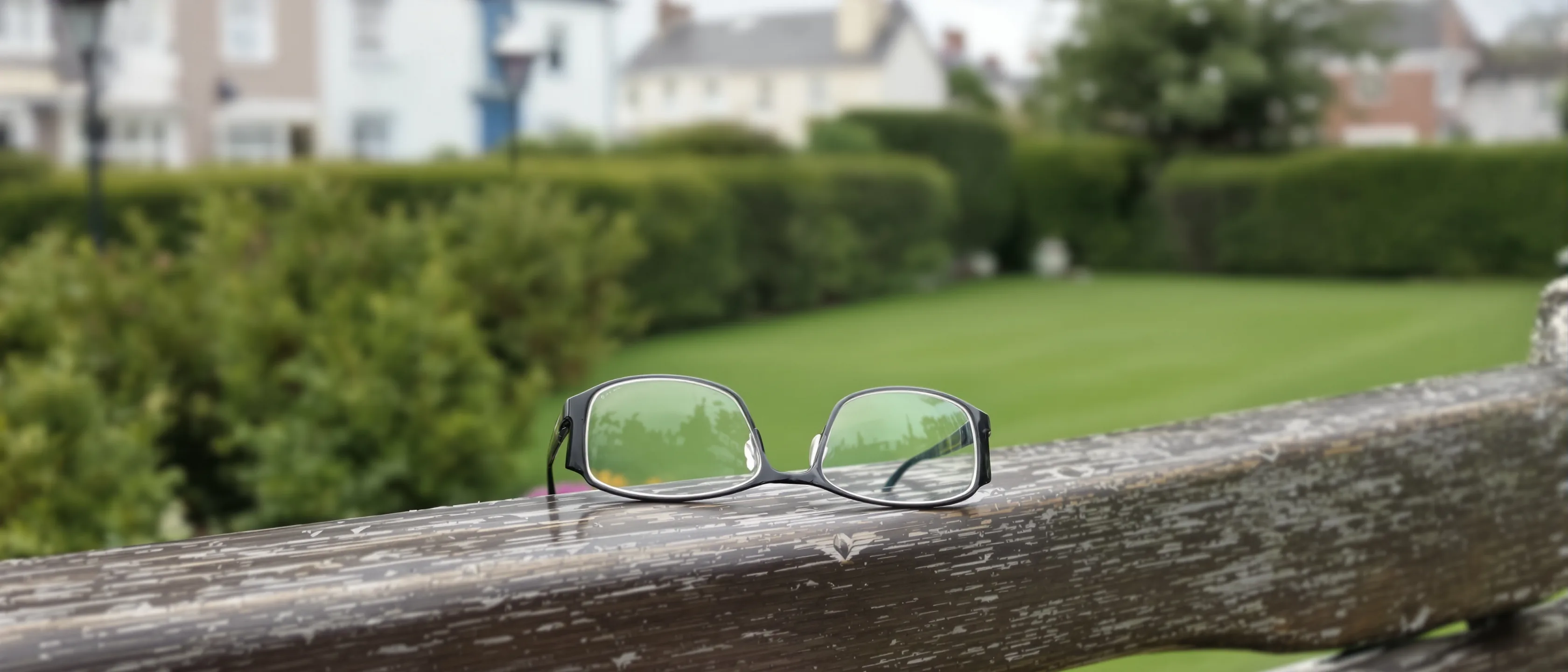 Eine Lesebrille liegt auf der Lehne einer verwitterten Gartenbank, dahinter unscharf Hecke und Rasen.