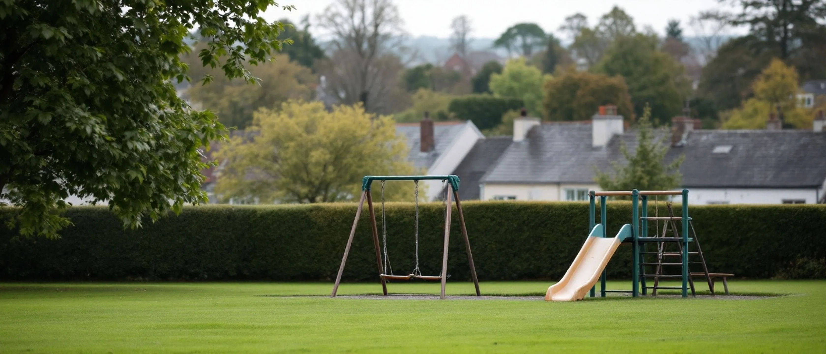Ein leerer Spielplatz hinter einer durchgehenden Hecke: Schaukeln und eine Rutsche, dahinter unscharf Schieferdächer und Bäume der Stadt.