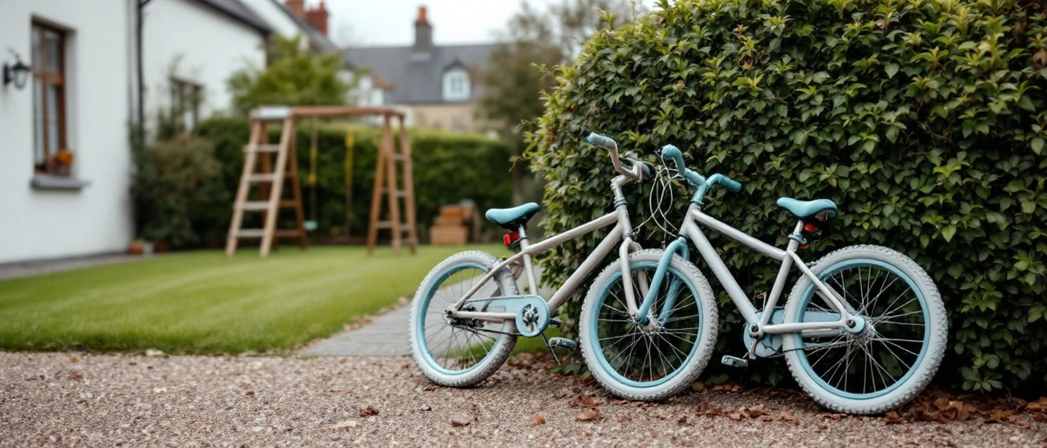 Zwei Kinderfahrräder lehnen an einer Gartenhecke neben einem Kiesweg, dahinter Rasen, ein Klettergerüst und eine Schaukel.
