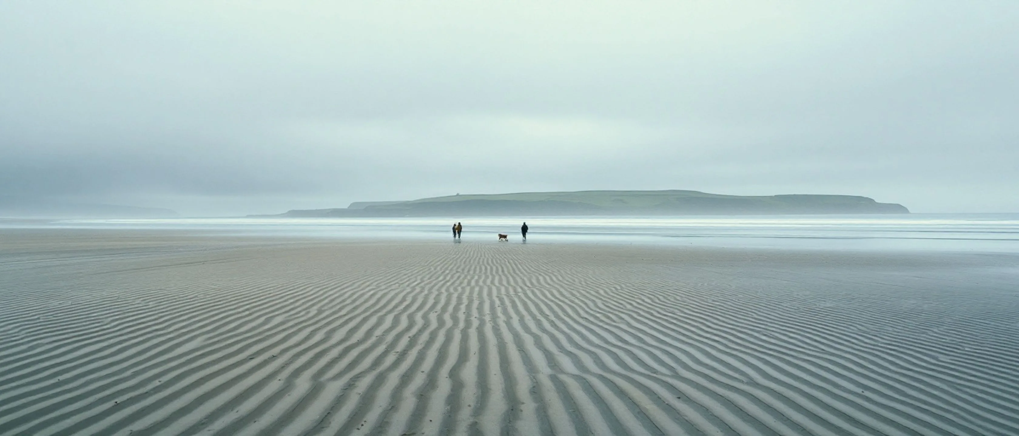 Ein sehr breiter Strand bei Niedrigwasser mit geriffeltem nassem Sand, zwei winzige Spaziergänger mit Hund in der Ferne, flache See.