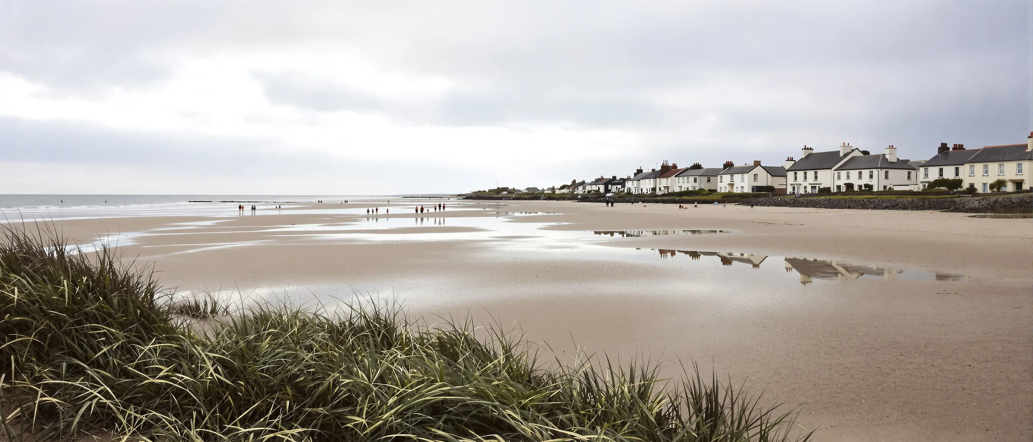 Sehr breiter Sandstrand bei Ebbe, Menschen winzig in der Ferne, nasser Sand spiegelt den hellen Wolkenhimmel, Dünengras vorn.