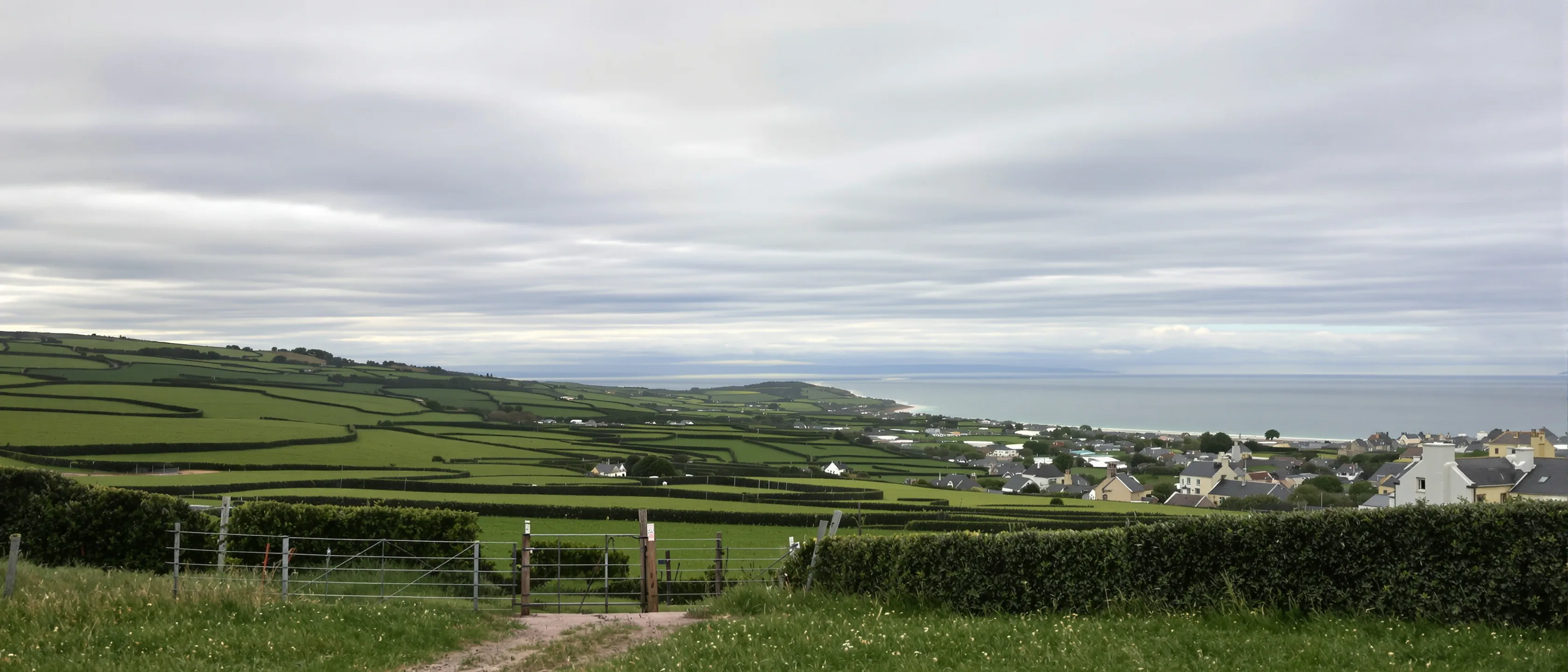 Blick von einem Hügel über Heckenfelder zum Meer, Wolkenschatten zeichnen Muster auf das Grün, ein Feldtor im Vordergrund.