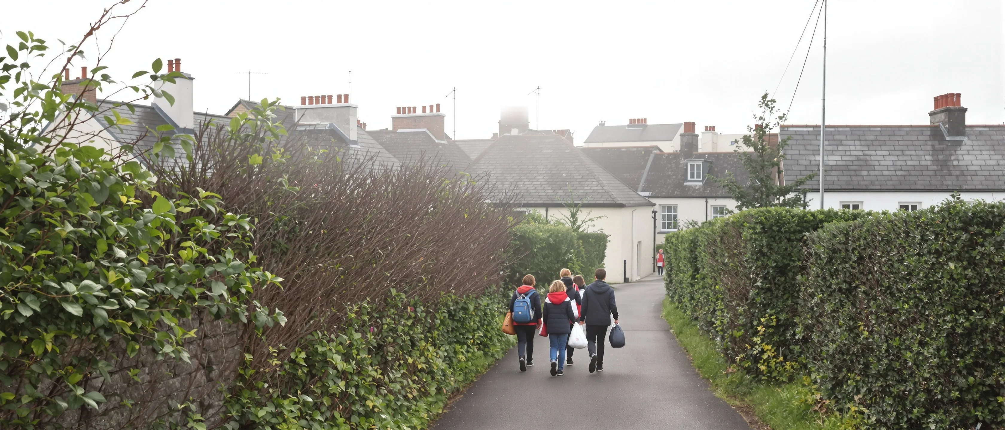 Eine kleine Gruppe Schulkinder mit Taschen von hinten auf einem Heckenweg zwischen Häusern, Morgenlicht.