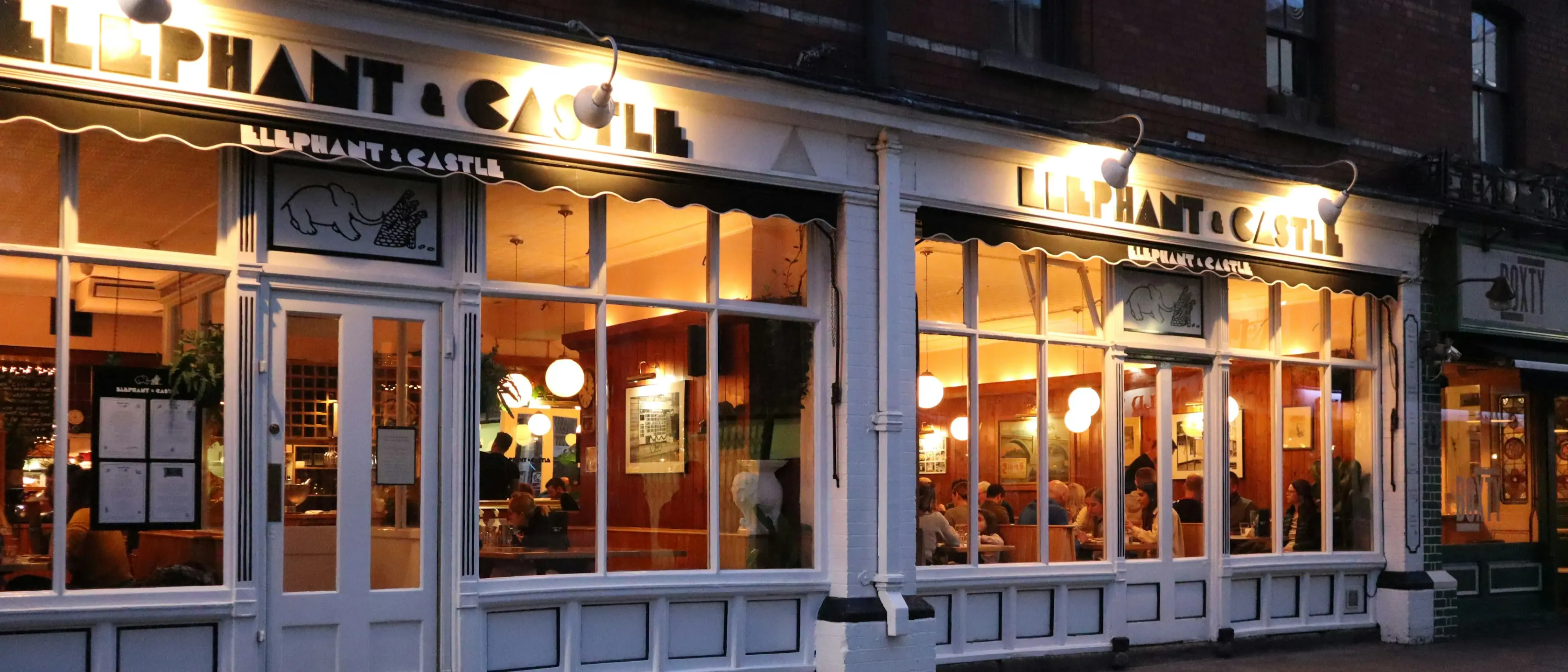 Ein beleuchtetes Restaurant mit großen Fenstern in einer Häuserzeile am Abend.