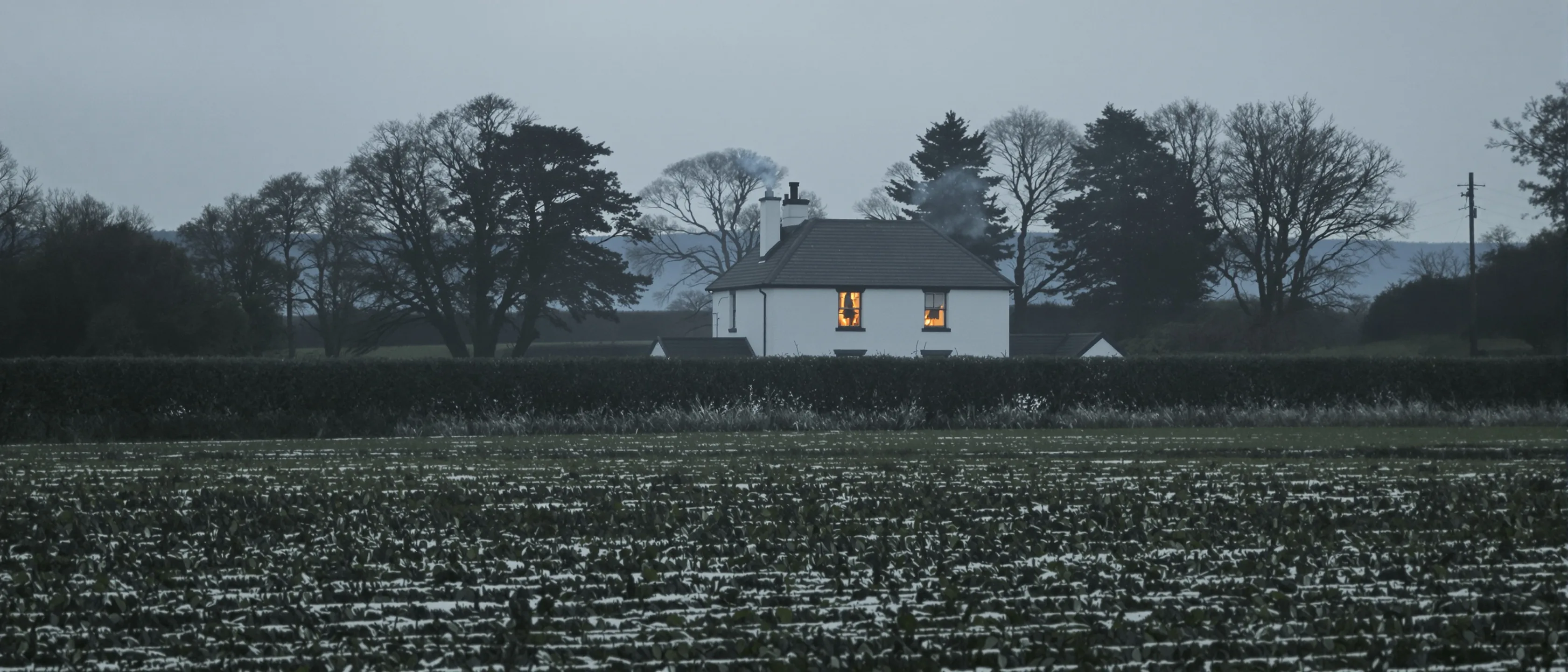 Einzelnes Bauernhaus in Winterfeldern mit einem erleuchteten Fenster und rauchendem Schornstein, dunkle Bäume dahinter.