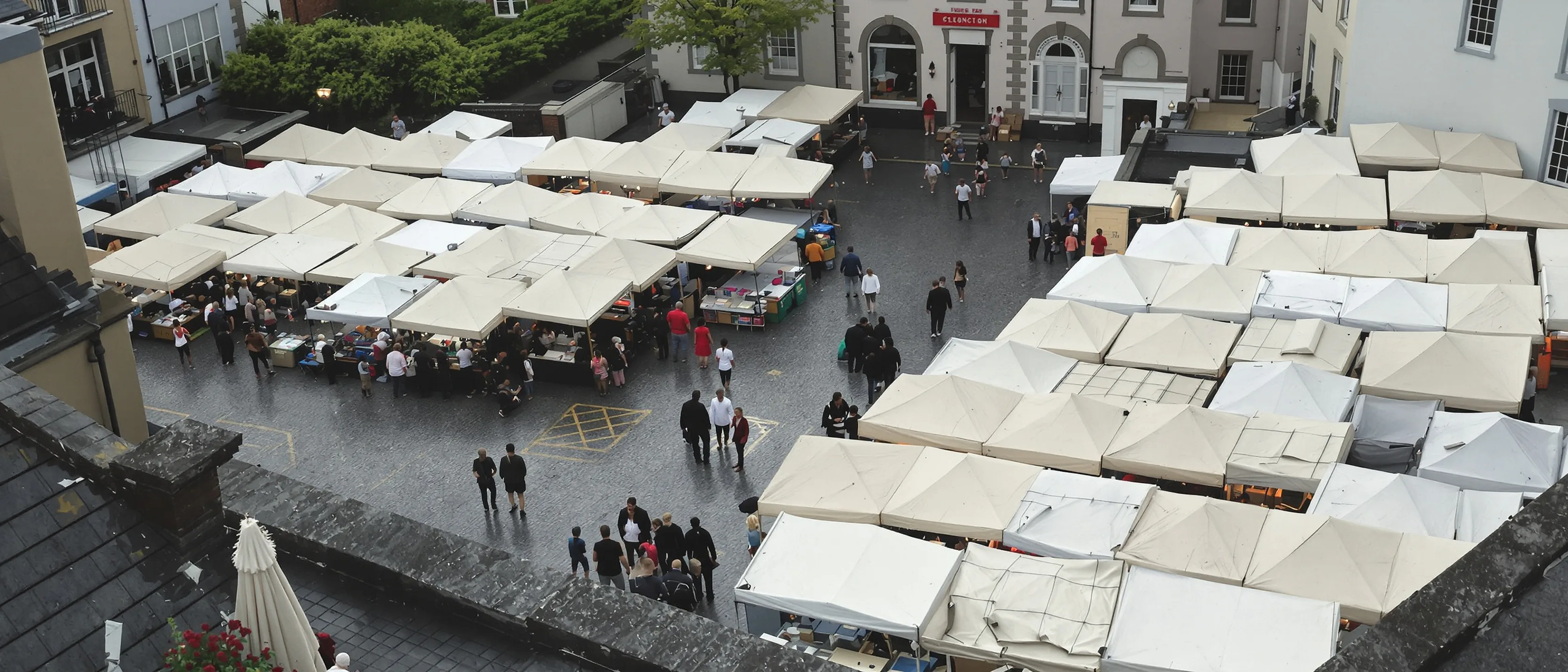 Marktstände mit hellen Planen auf einem Kleinstadtplatz, steil von oben gesehen, Menschen klein zwischen den Ständen.