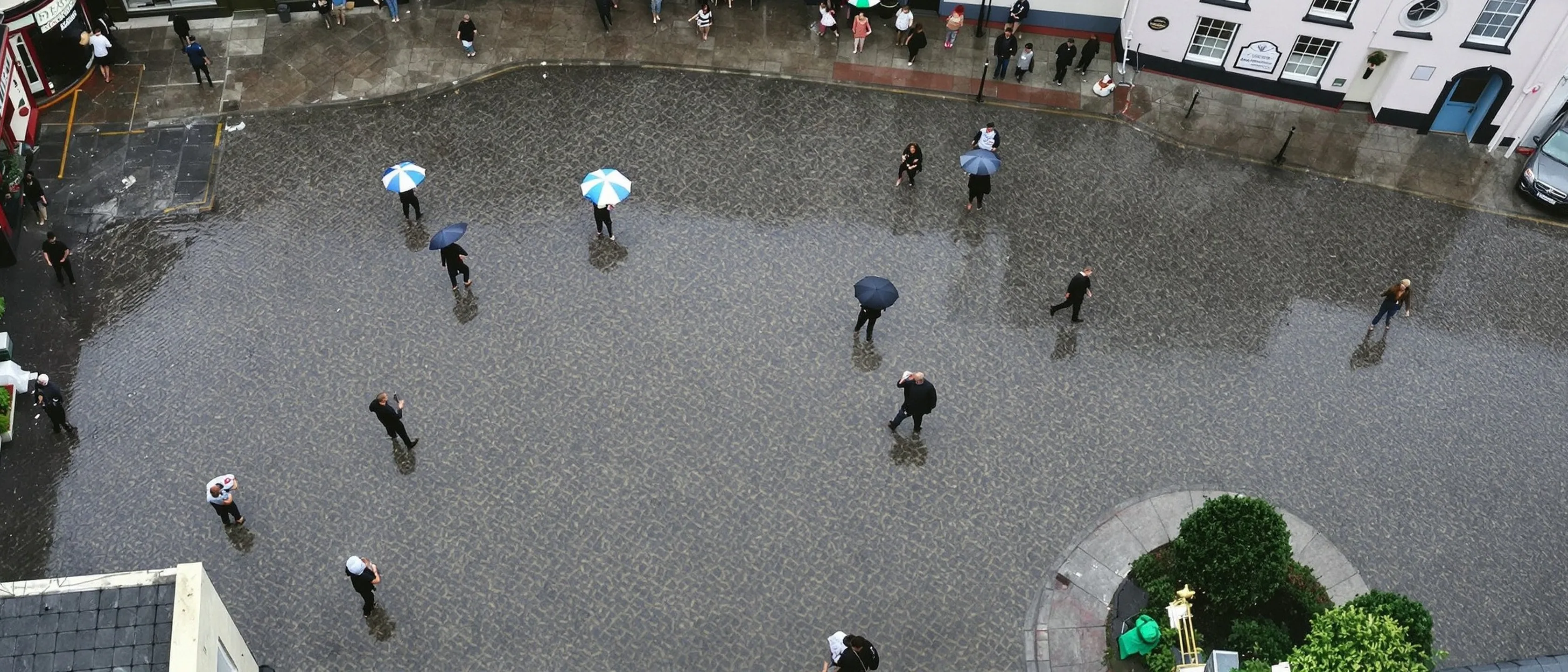 Ein regennasser Stadtplatz fast senkrecht von oben: Menschen mit Schirmen kreuzen ihn klein in verschiedene Richtungen.