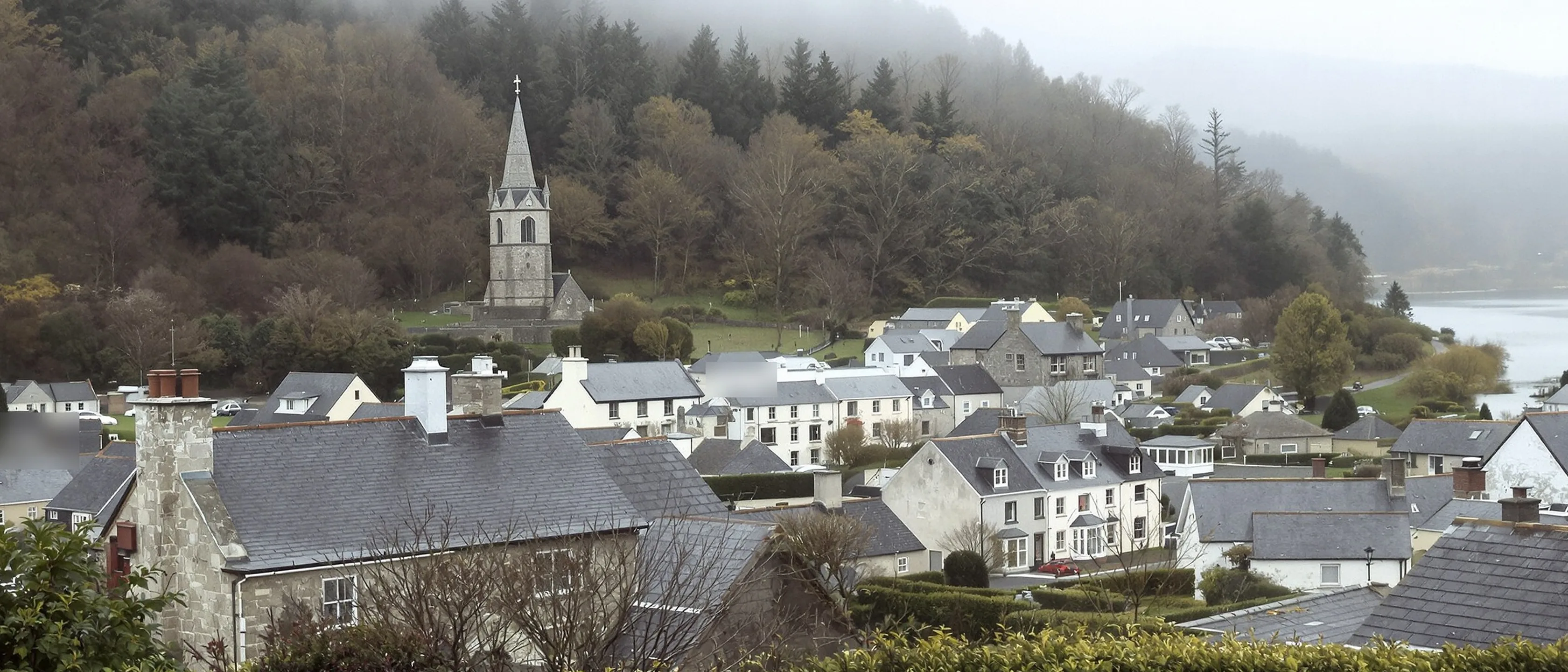 Ein kleiner Ort mit Schieferdächern und einem Kirchturm tief in einem bewaldeten Flusstal, Nebel hebt sich vom Wasser.