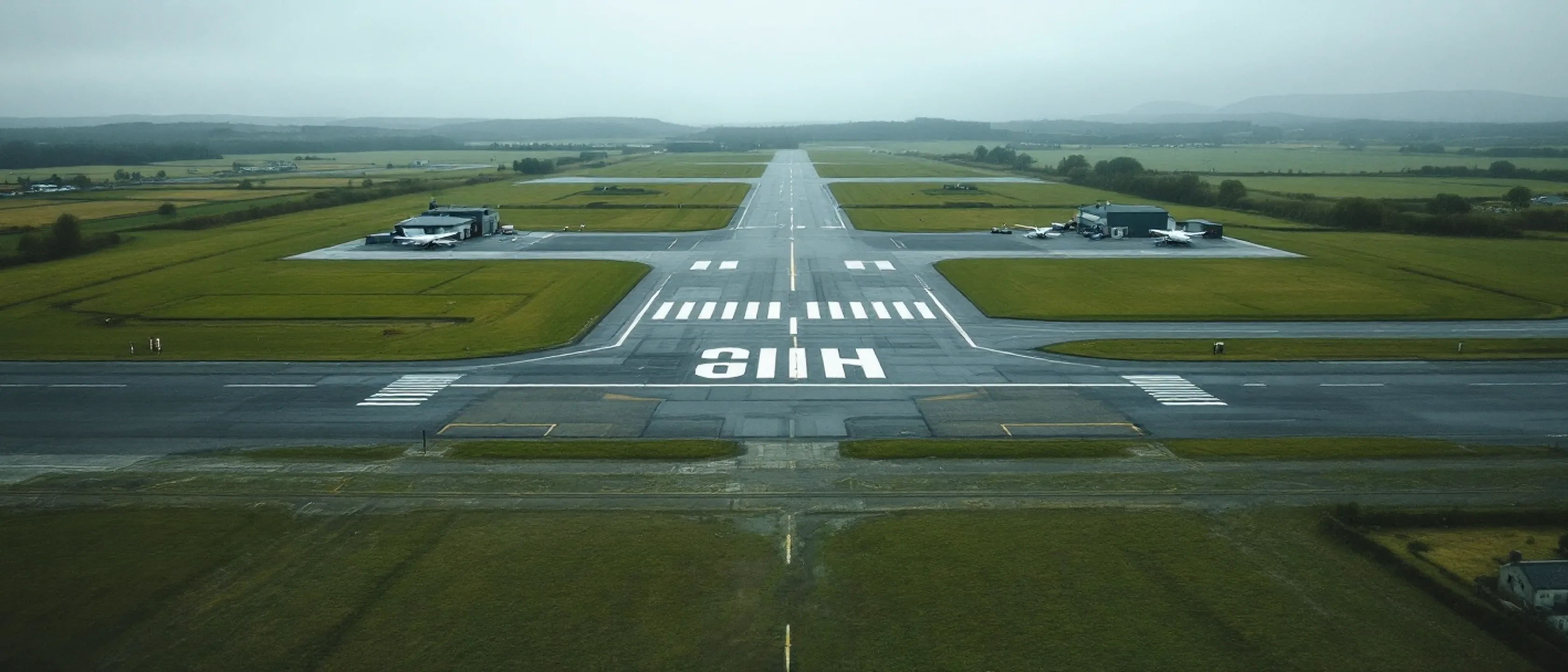 Ein kleines Regionalflugfeld aus der Ferne über eine Wiese gesehen, niedrige Gebäude, flaches Licht.