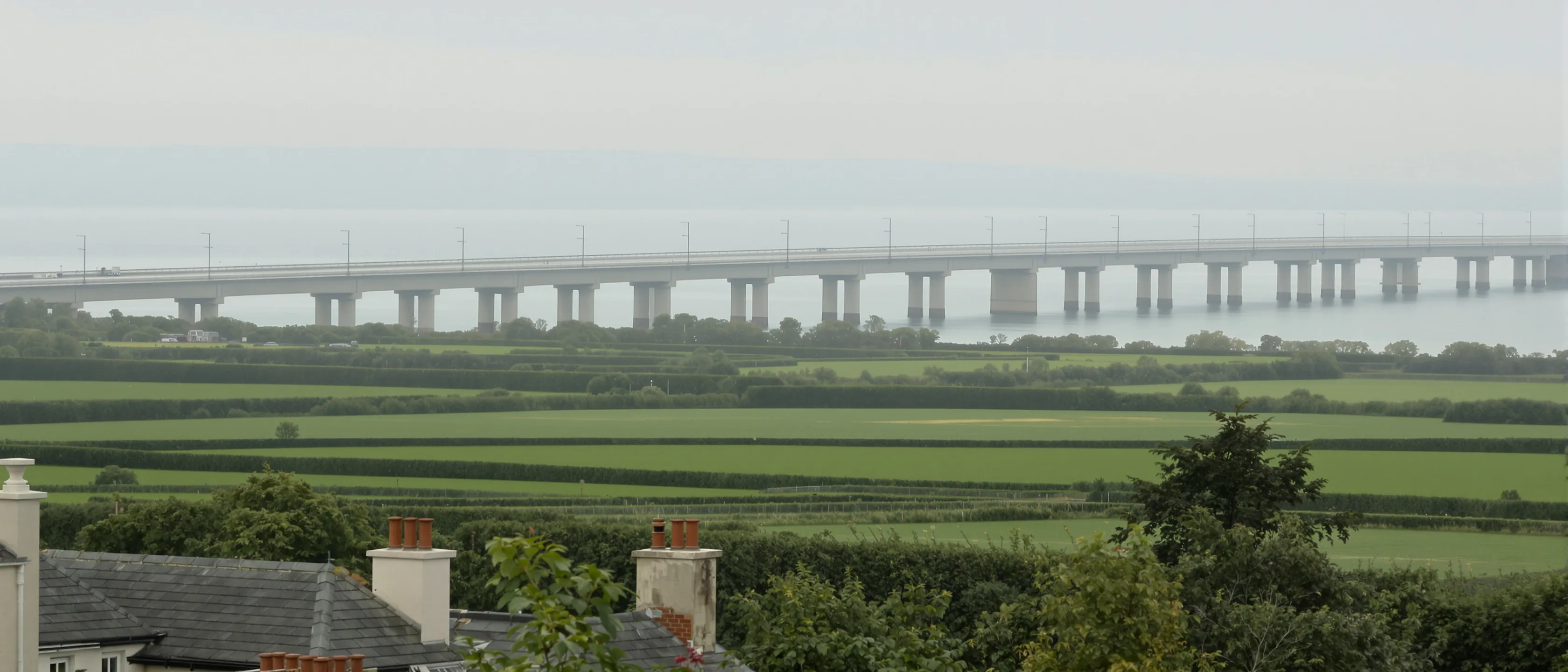 Eine lange moderne Brücke quert in der Ferne eine weite Flussmündung, davor Heckenfelder und Schornsteine im Dunst.