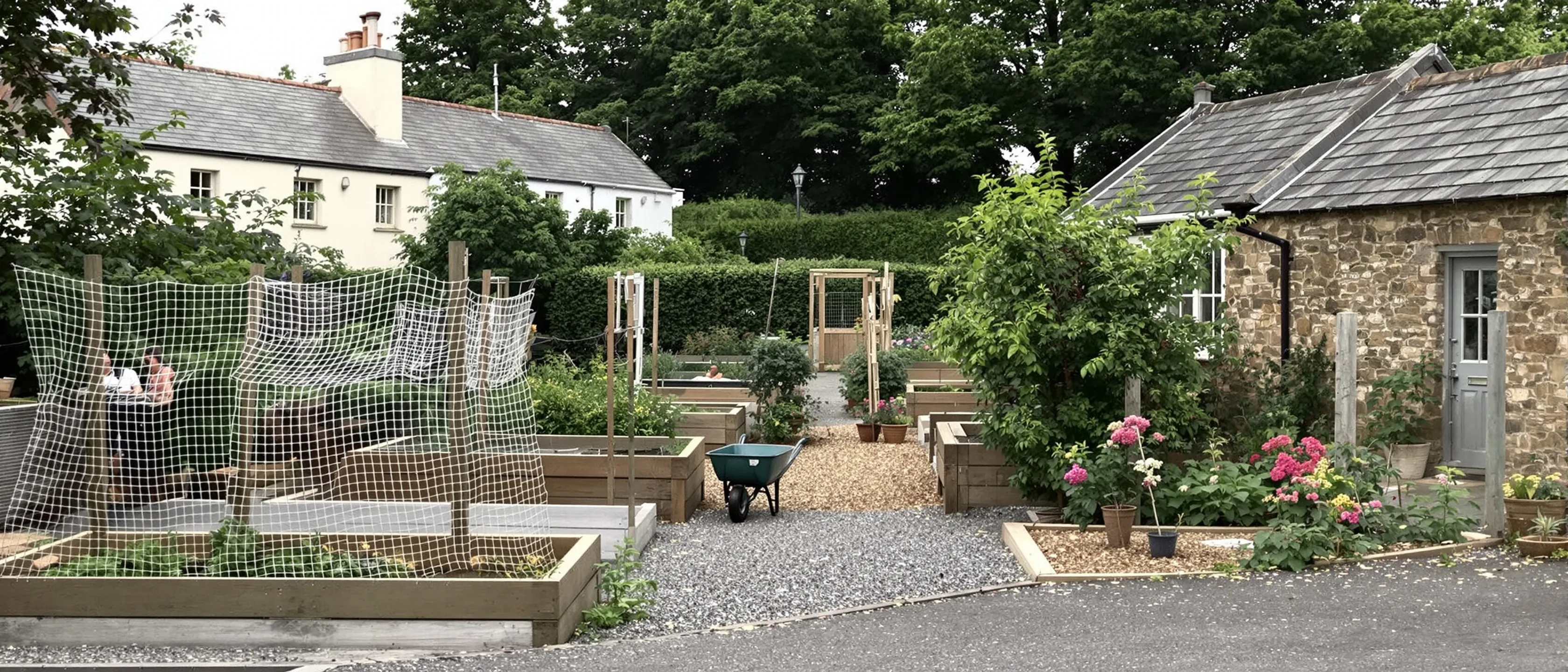 Ein Nutzgarten mit Hochbeeten und Obstnetzen zwischen Steinhäusern, eine Schubkarre auf dem Kiesweg.