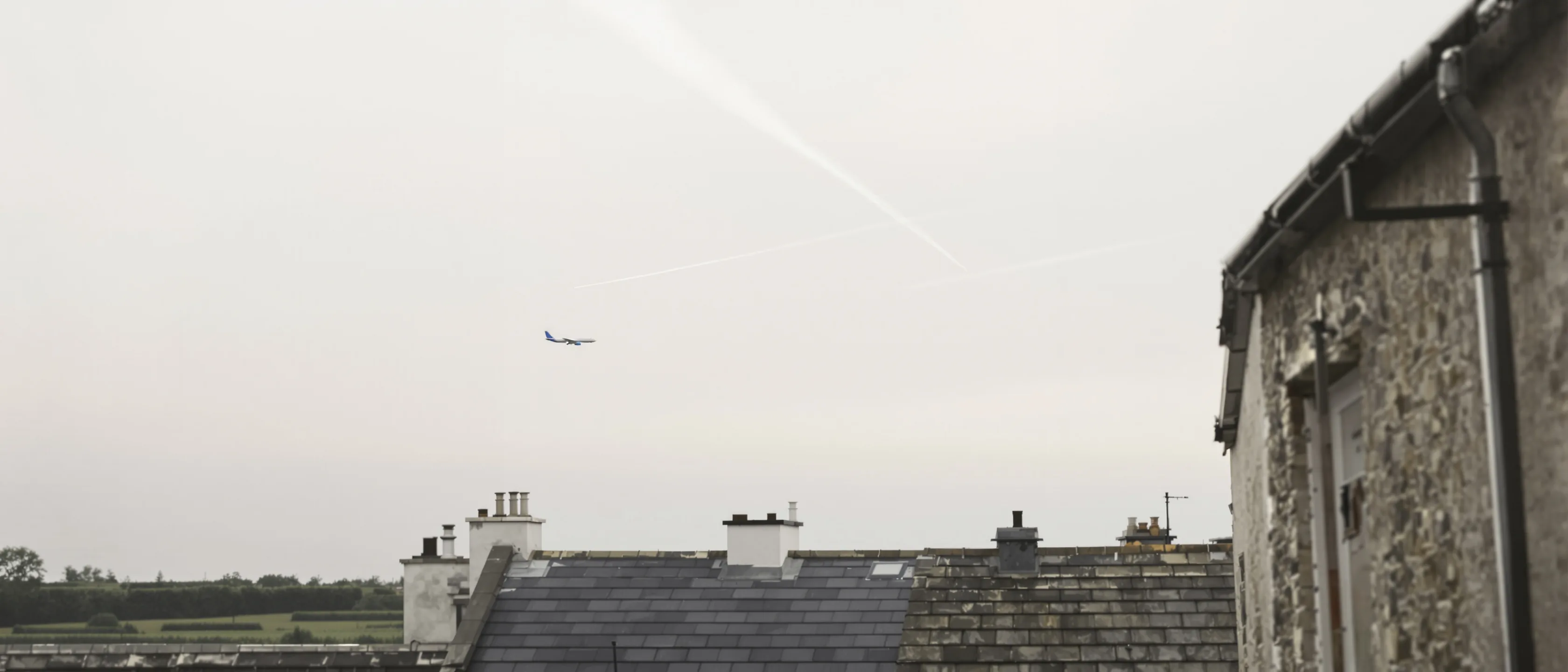 Ein Verkehrsflugzeug sehr hoch und klein mit dünnem Kondensstreifen über blassem Himmel, darunter Schornsteine und Dächer.